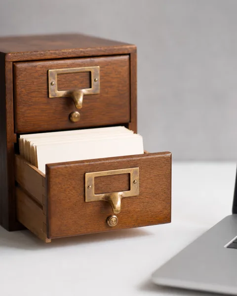 An open vintage wooden card catalog drawer with blank index cards beside a laptop